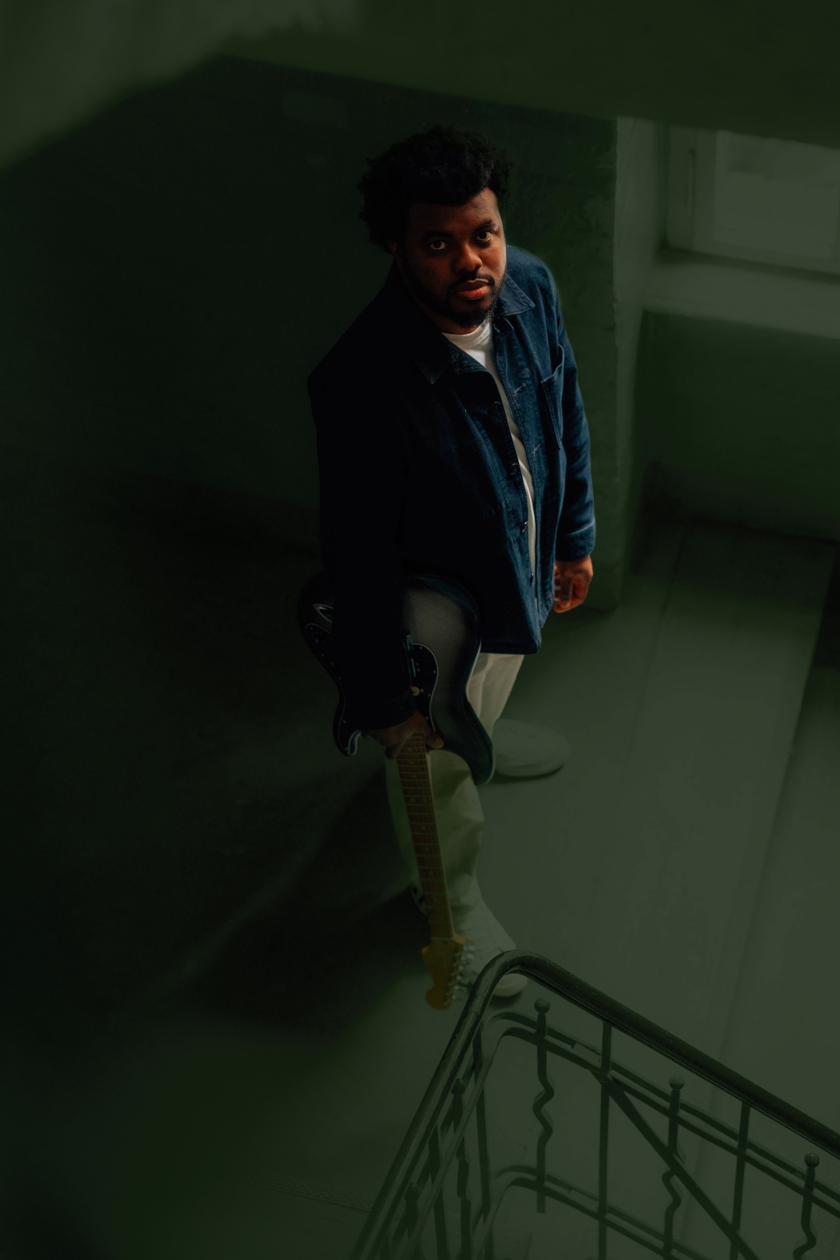 Diazno holding an electric guitar in a stairwell, looking up toward the camera